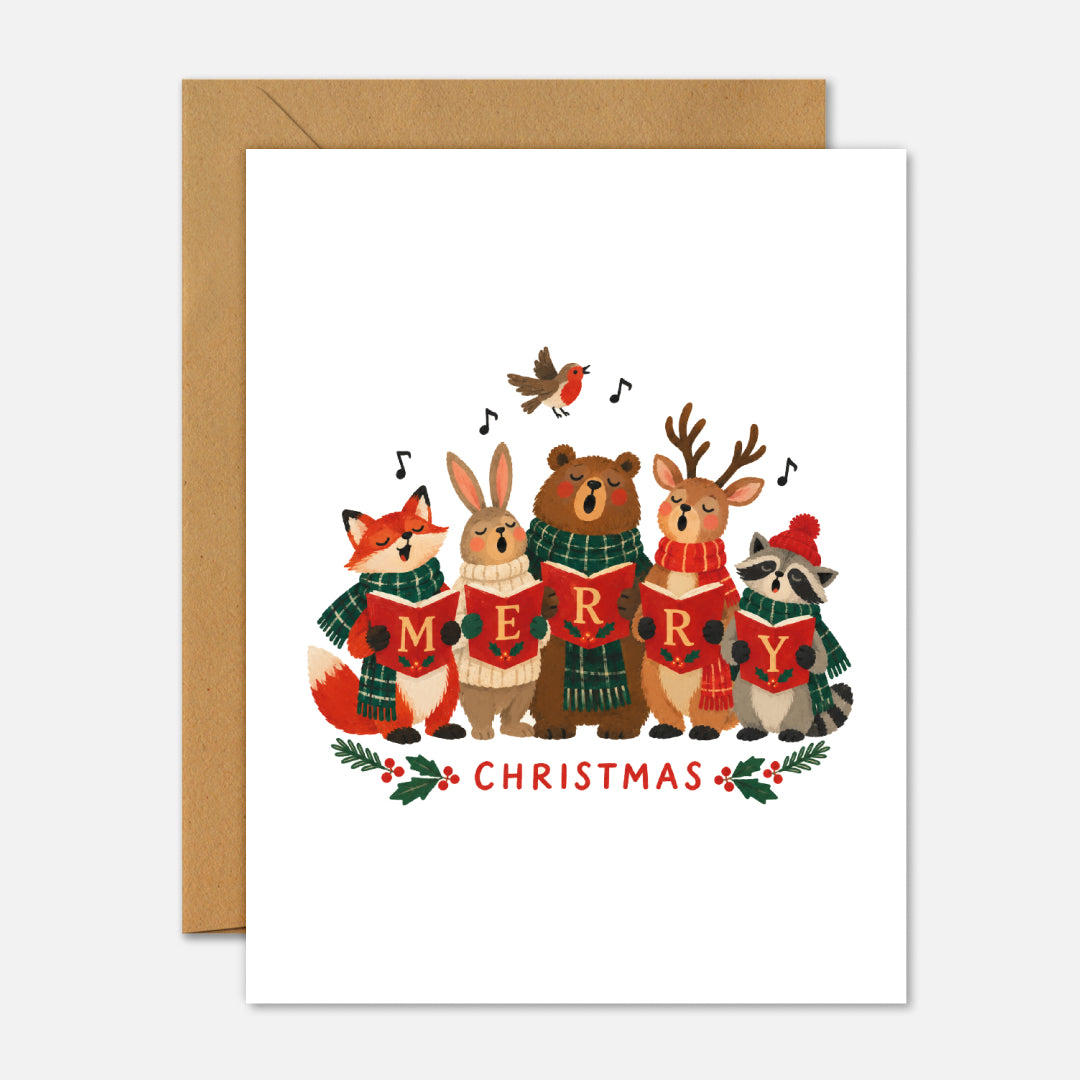 A white Christmas greeting card with a kraft envelope behind it. The card features an illustration of six woodland animals singing together under a robin, with the word 'CHRISTMAS' written below in red.