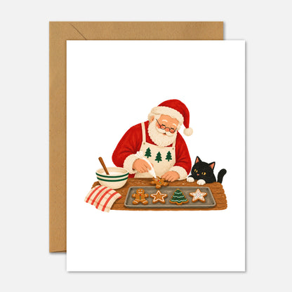 A Christmas greeting card featuring an illustration of Santa Claus baking cookies on a table with a black cat beside him, shown with a brown kraft envelope on a light gray background.