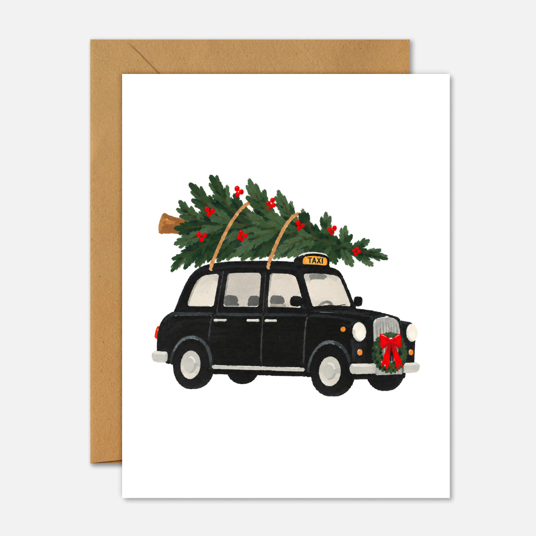 A white greeting card with a brown kraft envelope, featuring an illustration of a black London taxi carrying a Christmas tree on its roof.