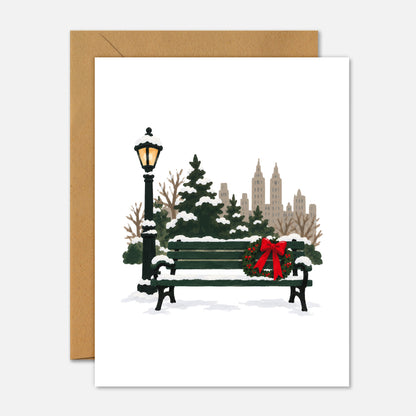 A white greeting card with a brown kraft envelope behind it, featuring an illustrated winter city scene with a bench, snow, trees, a lamppost, and a red bow.