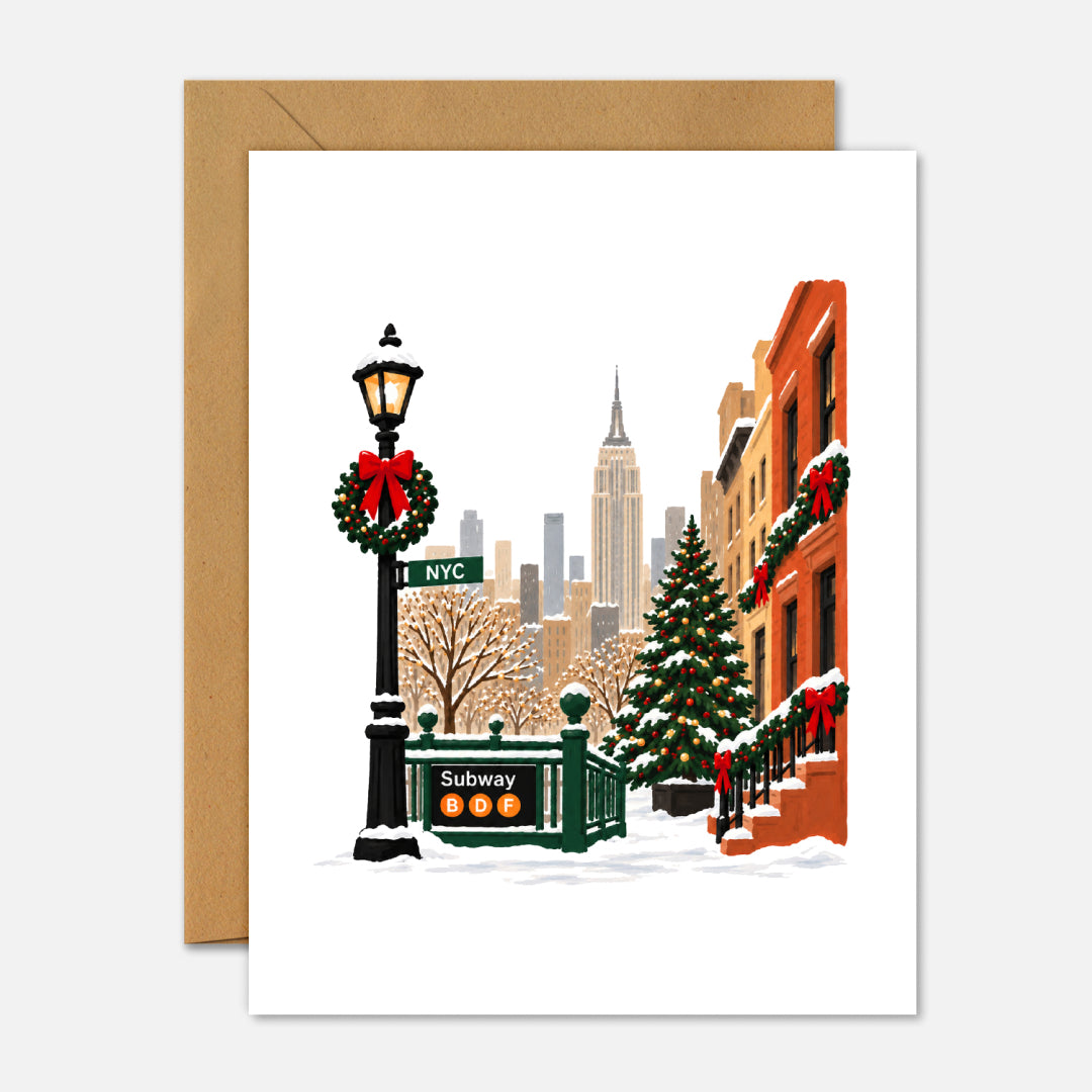 A greeting card with an illustrated New York City winter street scene, shown with a kraft envelope behind it on a light gray background. The card artwork includes snow, a lamppost with a red bow, a Christmas tree, a subway entrance, buildings, and the Empire State Building.