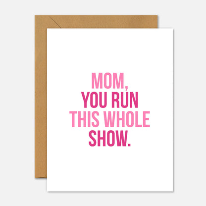 Greeting card with pink text 'Mom, you run this whole show.' on a white background with a brown envelope.