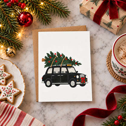 A Christmas-themed greeting card featuring an illustration of a black London taxi carrying a Christmas tree, styled on a marble surface with holiday decorations including pine branches, ornaments, wrapped gift boxes, cookies, and a mug of hot cocoa.