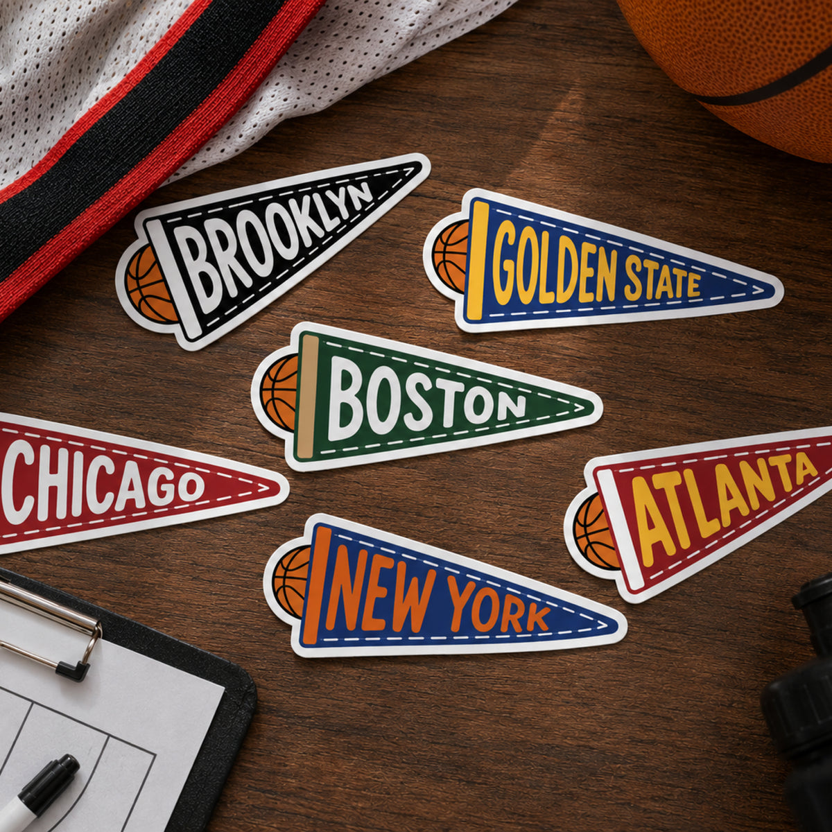 Basketball-themed city pennant stickers arranged on a wooden desk, with partial sports props including a basketball, a jersey, a clipboard, and binoculars around them.