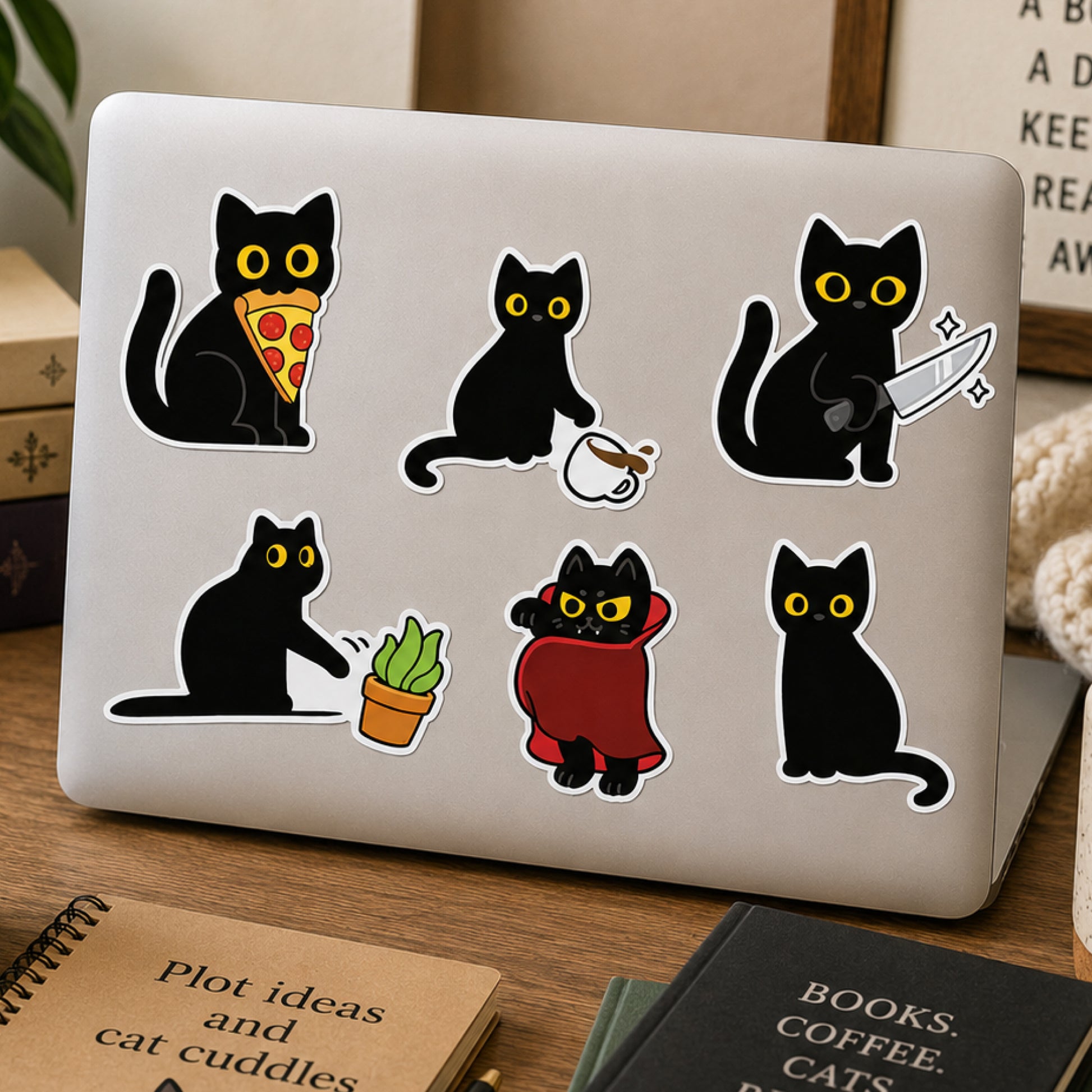 Black cat stickers with various whimsical designs are arranged on the lid of a silver laptop. The laptop sits on a wooden desk surrounded by books and a plant.