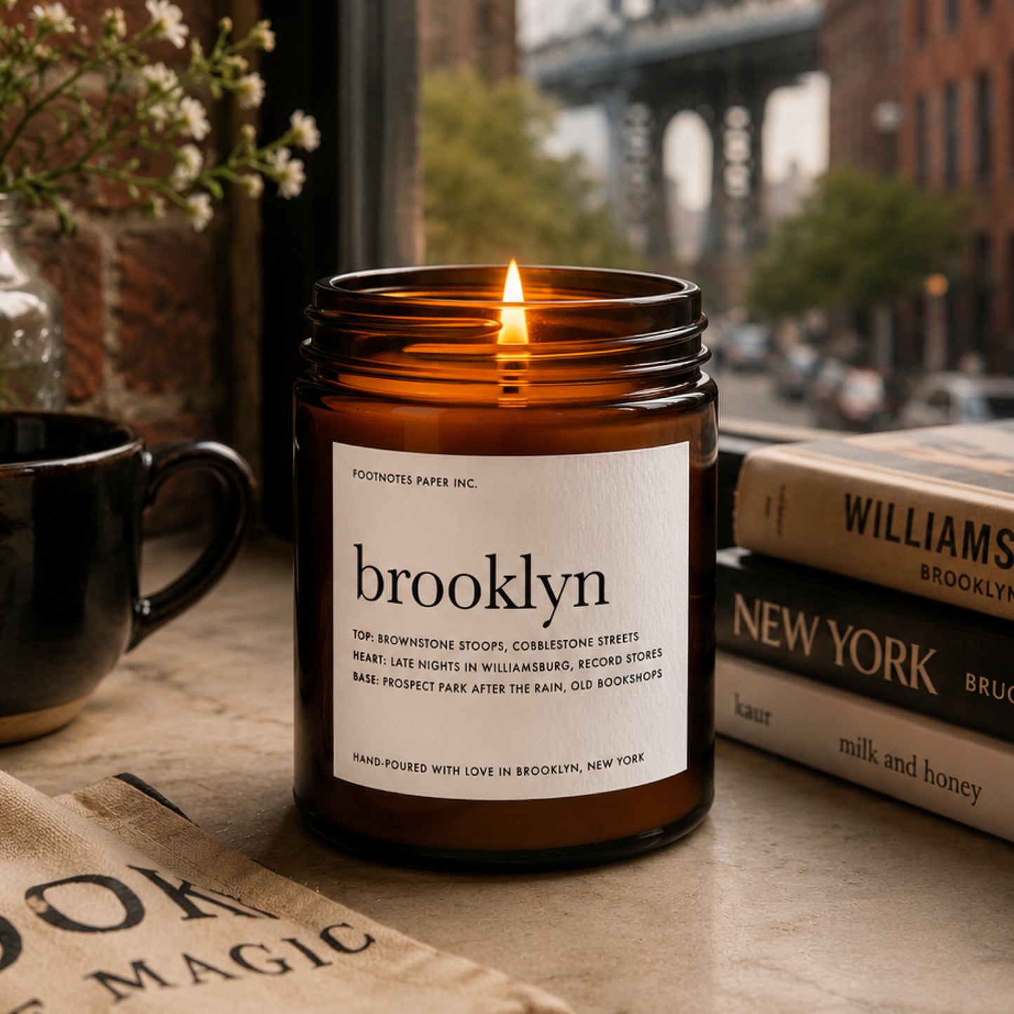 An amber glass jar candle with a white label reading 'brooklyn' and 'FOOTNOTES PAPER INC.' is lit on a tabletop.