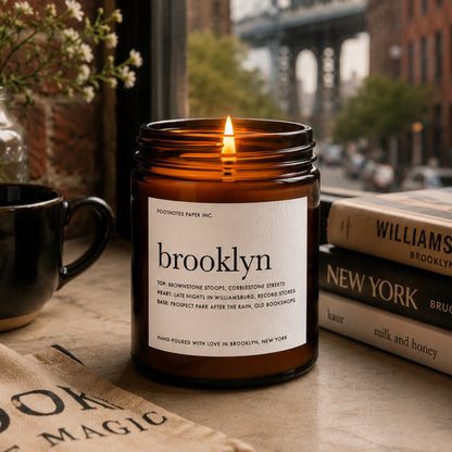 An amber glass jar candle with a white label reading 'brooklyn' and 'FOOTNOTES PAPER INC.' is lit on a tabletop.