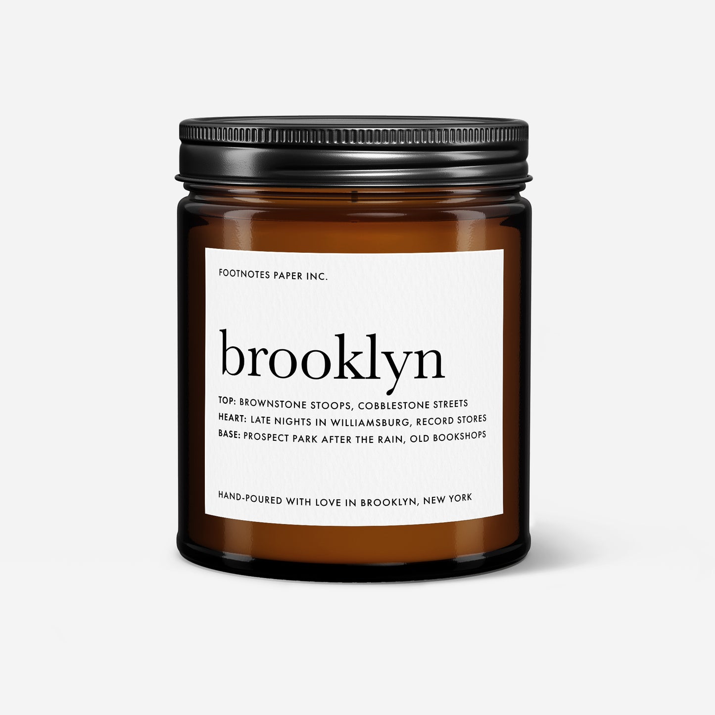 An amber glass jar candle with a black metal screw-on lid and a white paper label reading 'brooklyn' and 'FOOTNOTES PAPER INC.', photographed on a clean light gray background.