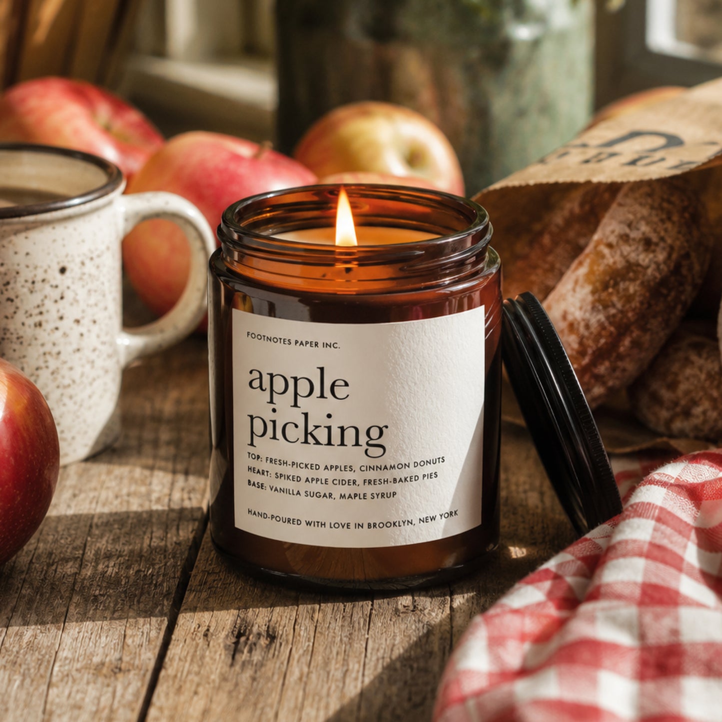 A lit amber glass jar candle labeled 'apple picking' by Posh Notes Paper Inc., set on a rustic wooden table with apples, a ceramic mug, and bread in a warm, cozy setting.
