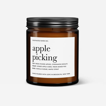An amber glass jar candle with a black metal lid and a white label reading 'apple picking' by Footnotes Paper Inc., shown on a light gray background.