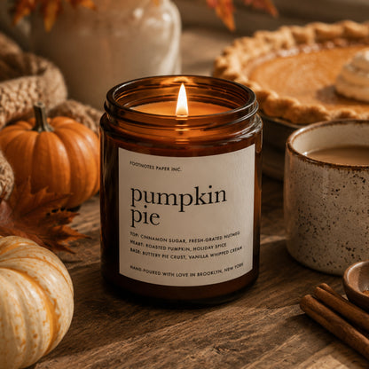 A lit amber glass candle jar labeled 'pumpkin pie' by Footnotes Paper Inc., styled on a wooden table with pumpkins, a pie, cinnamon sticks, and a mug in a warm autumn setting.