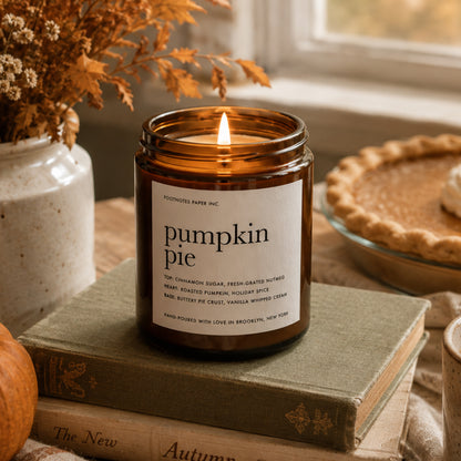 A lit amber glass candle jar labeled 'pumpkin pie' sits on stacked vintage-style books, with dried flowers, a pumpkin, and a pie in the background near a window.