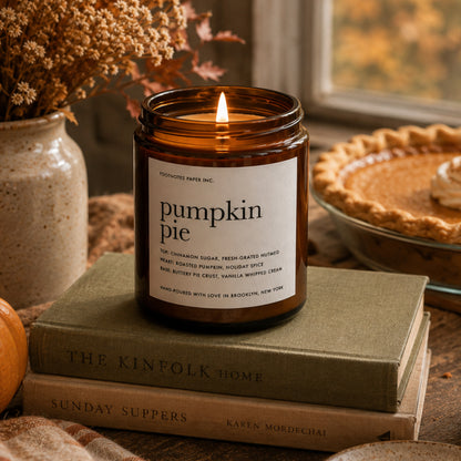 An amber glass jar candle with a lit flame sits on stacked books in a cozy autumn setting, with dried flowers in a vase and a pie in the background. The label reads 'pumpkin pie' and 'Pumpkin Pie Infused Paper Inc.