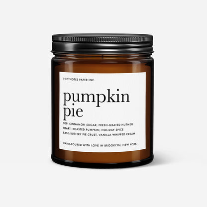 An amber glass jar candle with a black metal screw-on lid and a white paper label reading 'pumpkin pie' by Footnotes Paper Inc., shown on a light gray background.