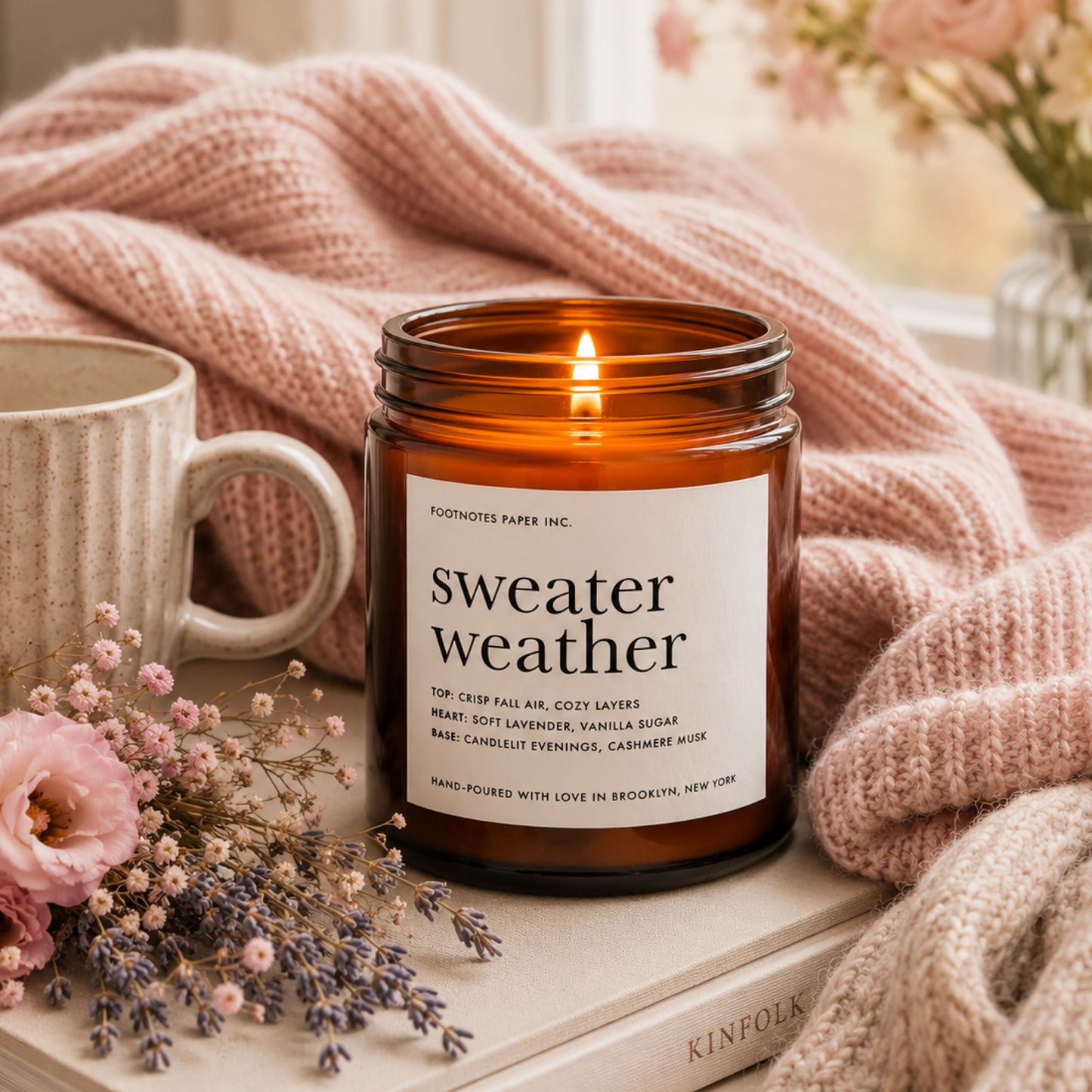 An amber glass jar candle labeled 'sweater weather' by Footnotes Paper Inc., lit and styled on a book with a pink knit blanket, a ceramic mug, and dried flowers.
