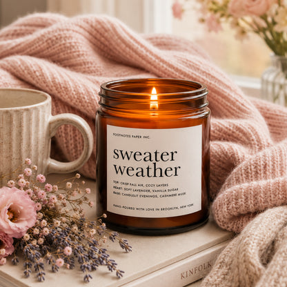 An amber glass jar candle labeled 'sweater weather' by Footnotes Paper Inc., lit and styled on a book with a pink knit blanket, a ceramic mug, and dried flowers.