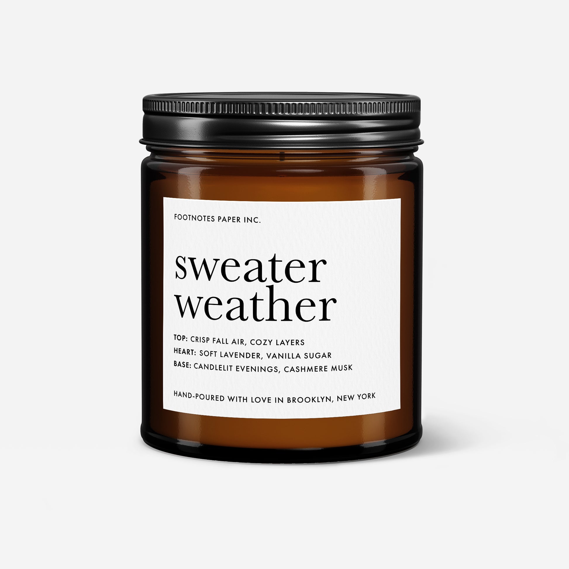 An amber glass jar candle with a black metal lid and a white label reading 'sweater weather' by Footnotes Paper Inc., shown on a light gray background.