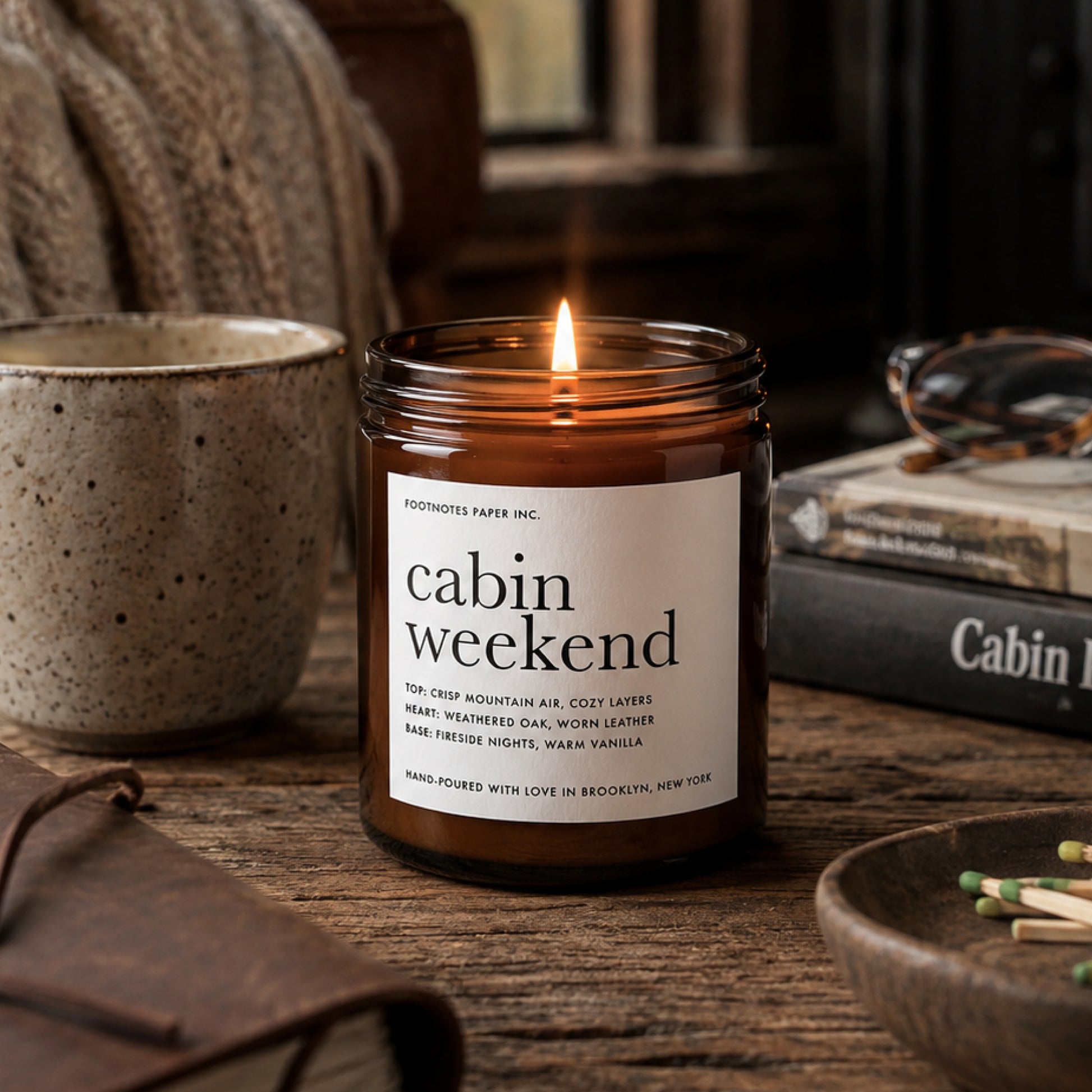 A lit amber glass jar candle labeled 'cabin weekend' by Footnotes Paper Inc., styled on a wooden table with a ceramic mug, books, glasses, and a leather pouch in a cozy indoor setting.