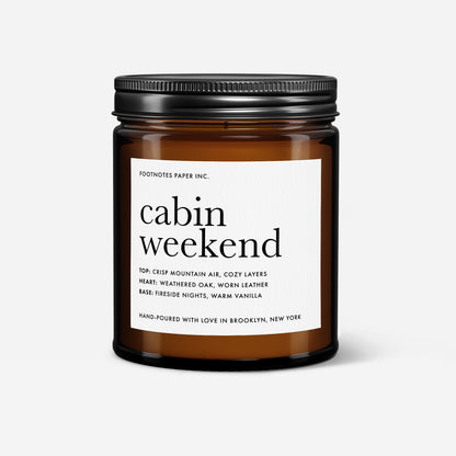An amber glass jar candle with a black metal screw-on lid and a white paper label reading 'cabin weekend' by Footnotes Paper Inc., shown on a light gray background.