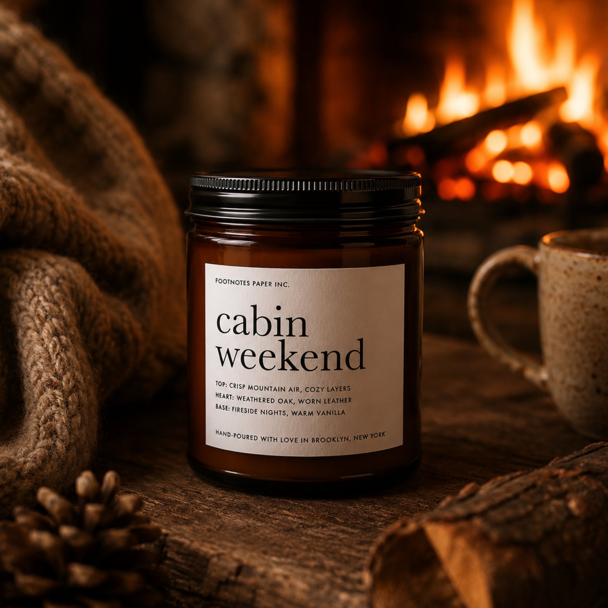 A candle jar labeled 'cabin weekend' by Footnotes Paper Inc. sits on a rustic wooden surface with a cozy fireplace, knitted blanket, pinecone, and mug in the background.