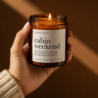 A hand holds a lit amber glass jar candle labeled 'cabin weekend' by Footnotes Paper Inc. against a warm brown background.