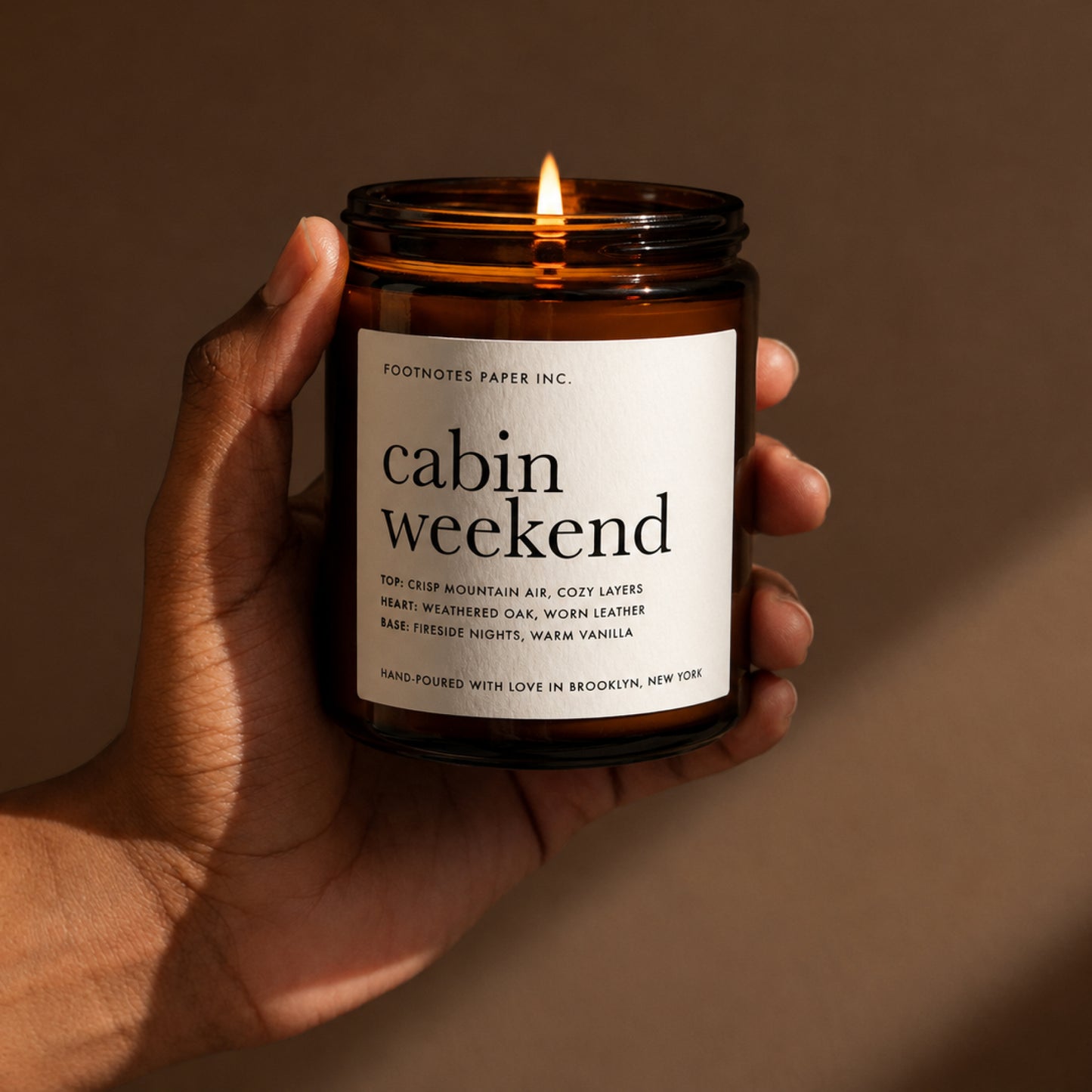 A hand holds a lit amber glass candle jar labeled 'cabin weekend' by Footnotes Paper Inc. against a warm brown background.