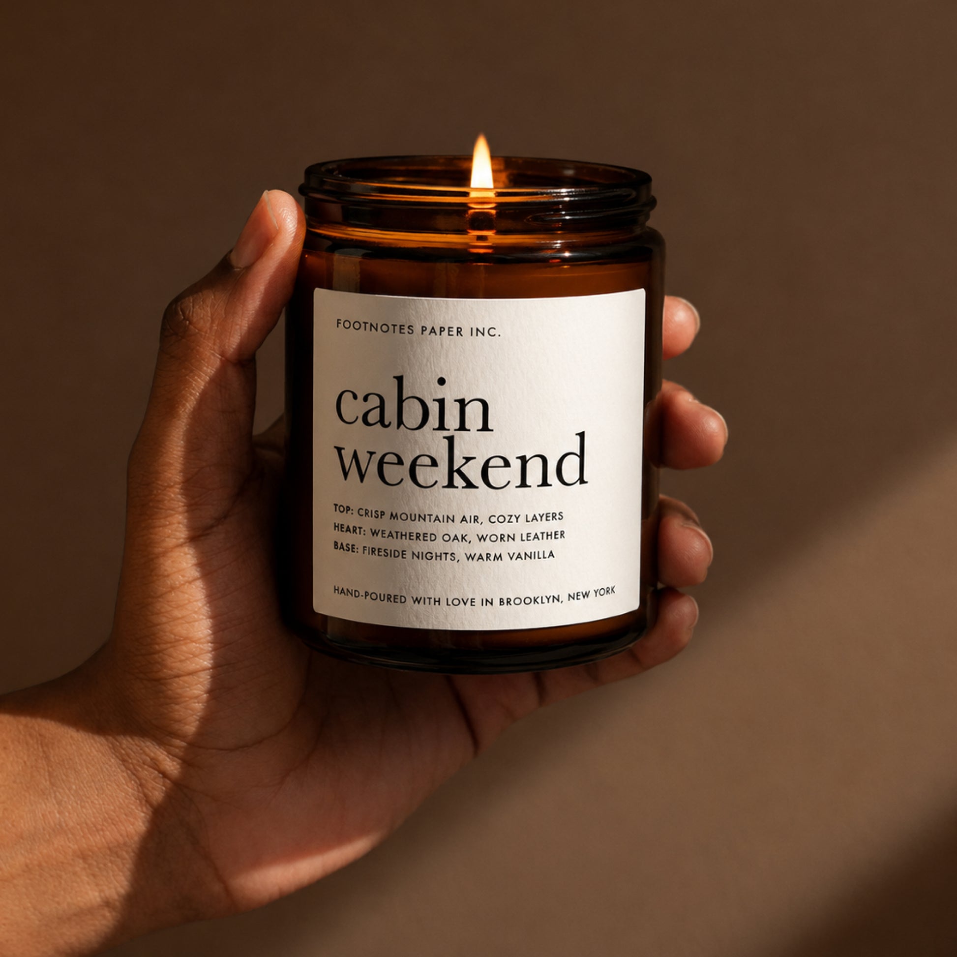 A hand holds a lit amber glass candle jar labeled 'cabin weekend' by Footnotes Paper Inc. against a warm brown background.