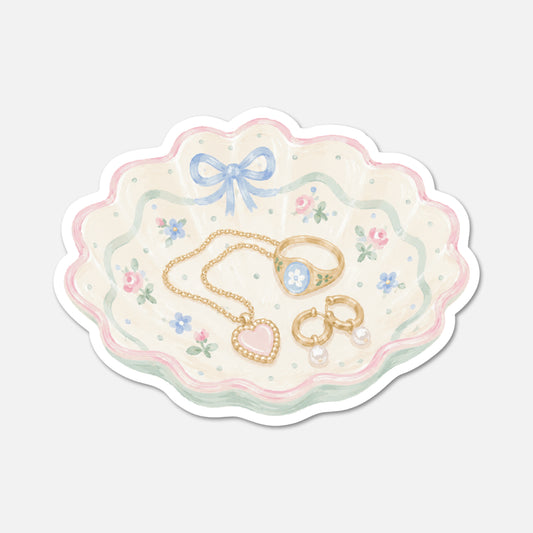 A scalloped ceramic jewelry tray with a pastel floral design, shown holding gold jewelry including a heart pendant necklace, a ring with a blue flower, and hoop earrings, on a light gray background.