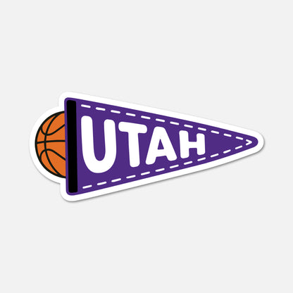 A die-cut sticker of a purple Utah basketball pennant with a partial orange basketball peeking out from behind it, on a light gray background.