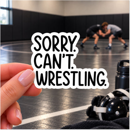 A hand holds a die-cut sticker with the text 'SORRY. CAN'T. WRESTLING.'