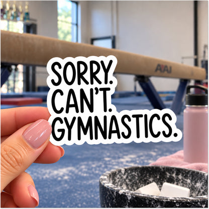 A hand with pink manicured nails holds a die-cut sticker that reads 'SORRY. CAN'T. GYMNASTICS.'