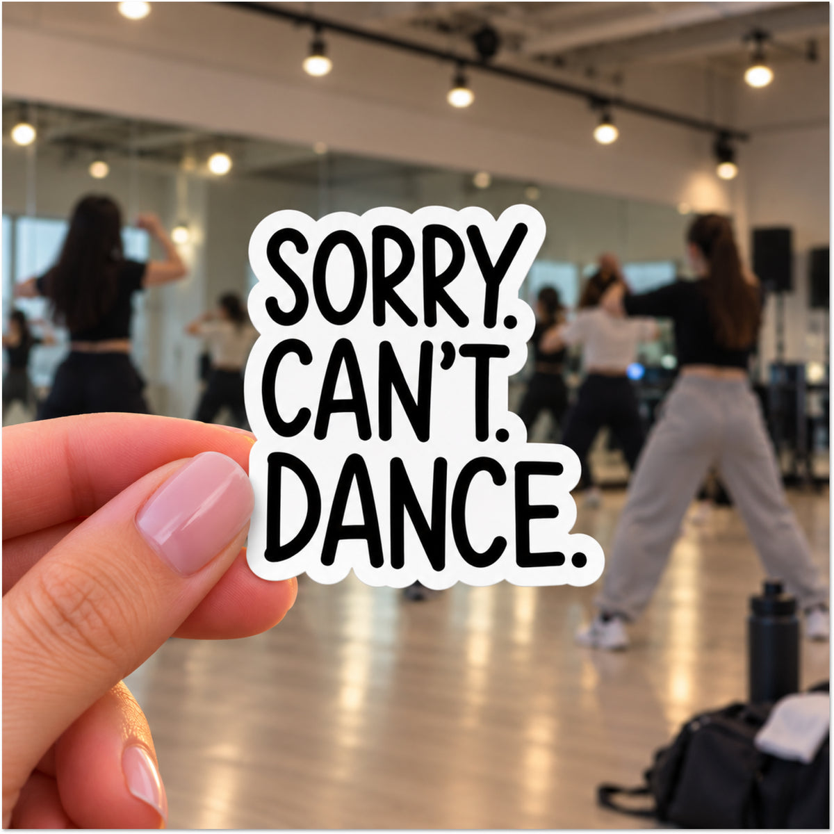 A hand with pink manicured nails holds a die-cut sticker in the foreground, with a blurred dance studio scene in the background where people are dancing. The sticker reads 'SORRY. CAN'T. DANCE.'