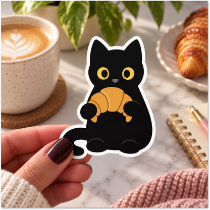 A die-cut sticker featuring a cute black cat holding a golden croissant, held by a hand with dark burgundy nail polish. The background is a cozy, softly blurred lifestyle scene with a cup of latte, a croissant on a plate, and a notebook on a marble surface.