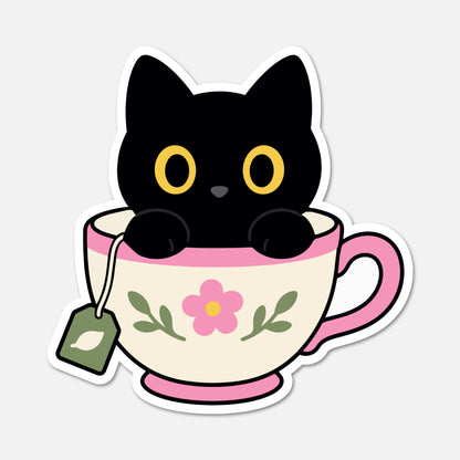 A die-cut sticker featuring a cute cartoon black cat with yellow eyes sitting inside a cream-colored teacup with pink trim and a pink flower on the front. A green tea bag hangs from the side.